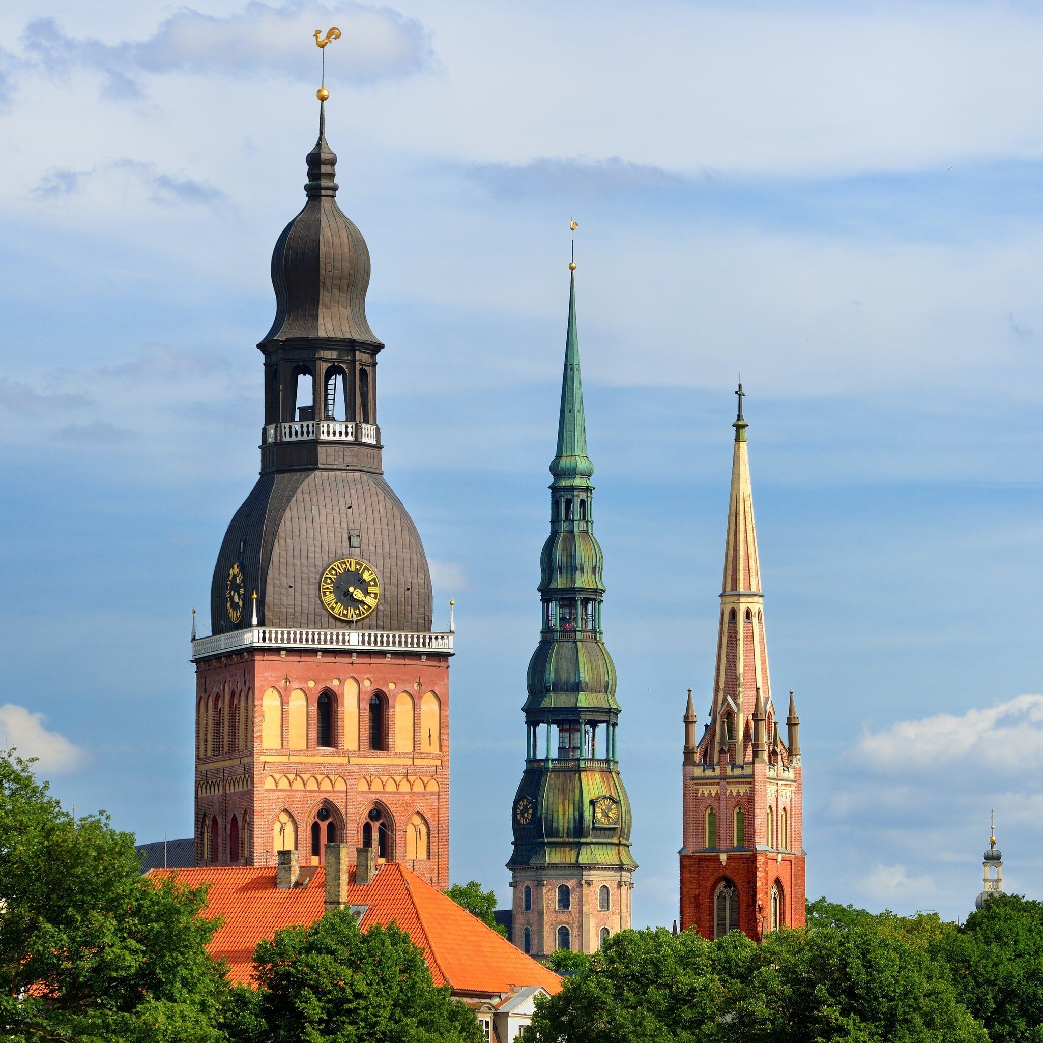 Three church towers above treetops: a domed clock tower, a tall tiered green spire and a slender red-brick spire