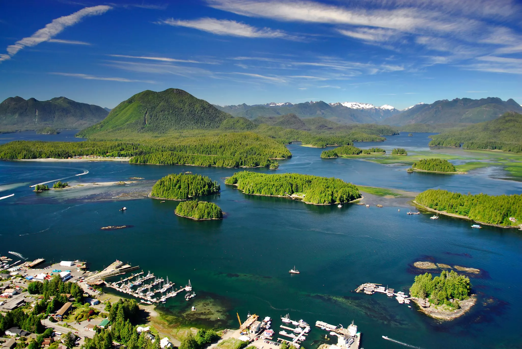 Aerial view of small, tree-covered islands with mountains in the distance and a harbor town in the foreground on a mosty sunny day.