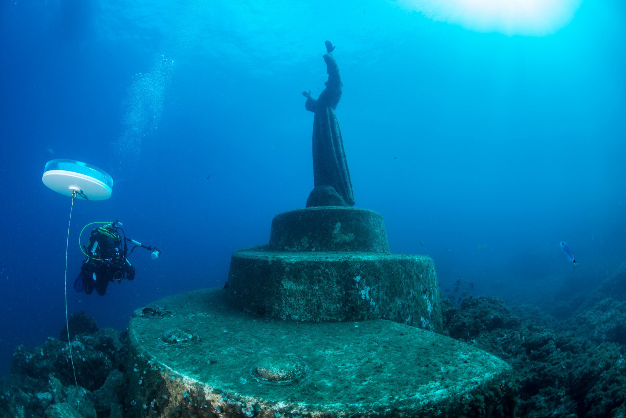 A scuba diver near an underwater statue of a man with his arms in the air reaching towards the sky.