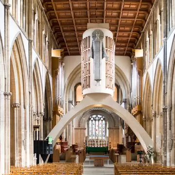 A pale aluminium figure of Christ mounted on a concrete drum spanning a Gothic nave on a parabolic arch, above rows of light wooden chairs.