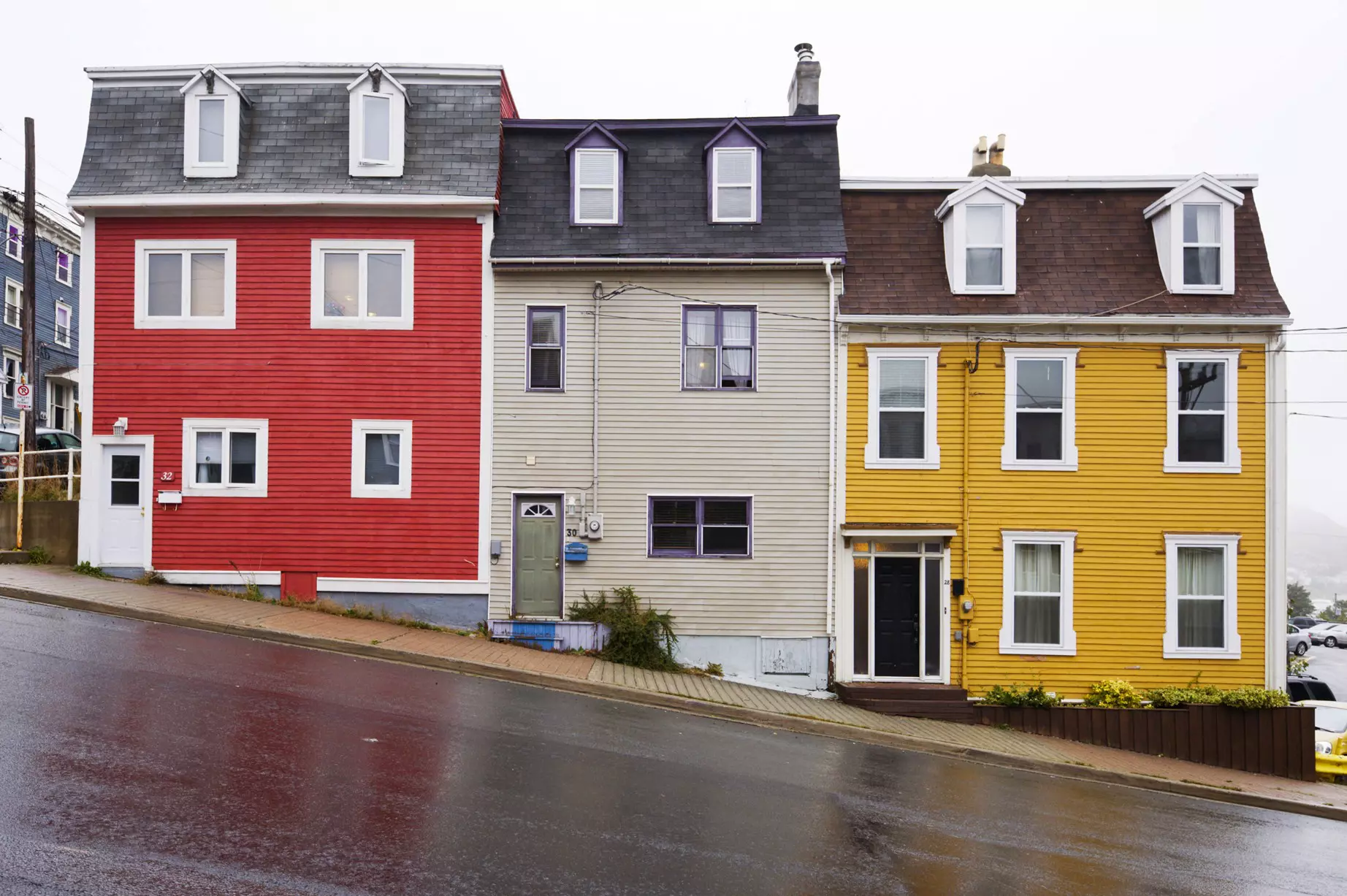 Three colorful houses in a row – one red, one cream and one yellow – on a rain-slicked, steeply sloped street.