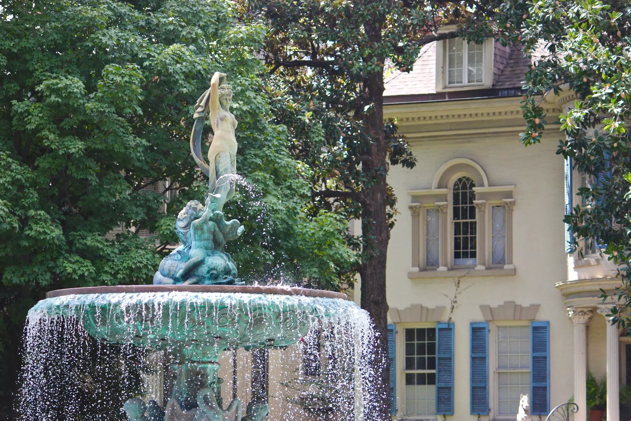 Close-up of water fountain with statuary atop and historic cream-colored home with blue shutters in the background.