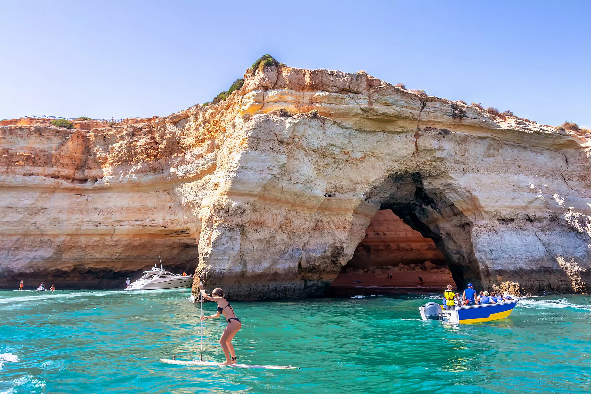 A paddleboarder and a small tour boat pass near the entrance to a cave.