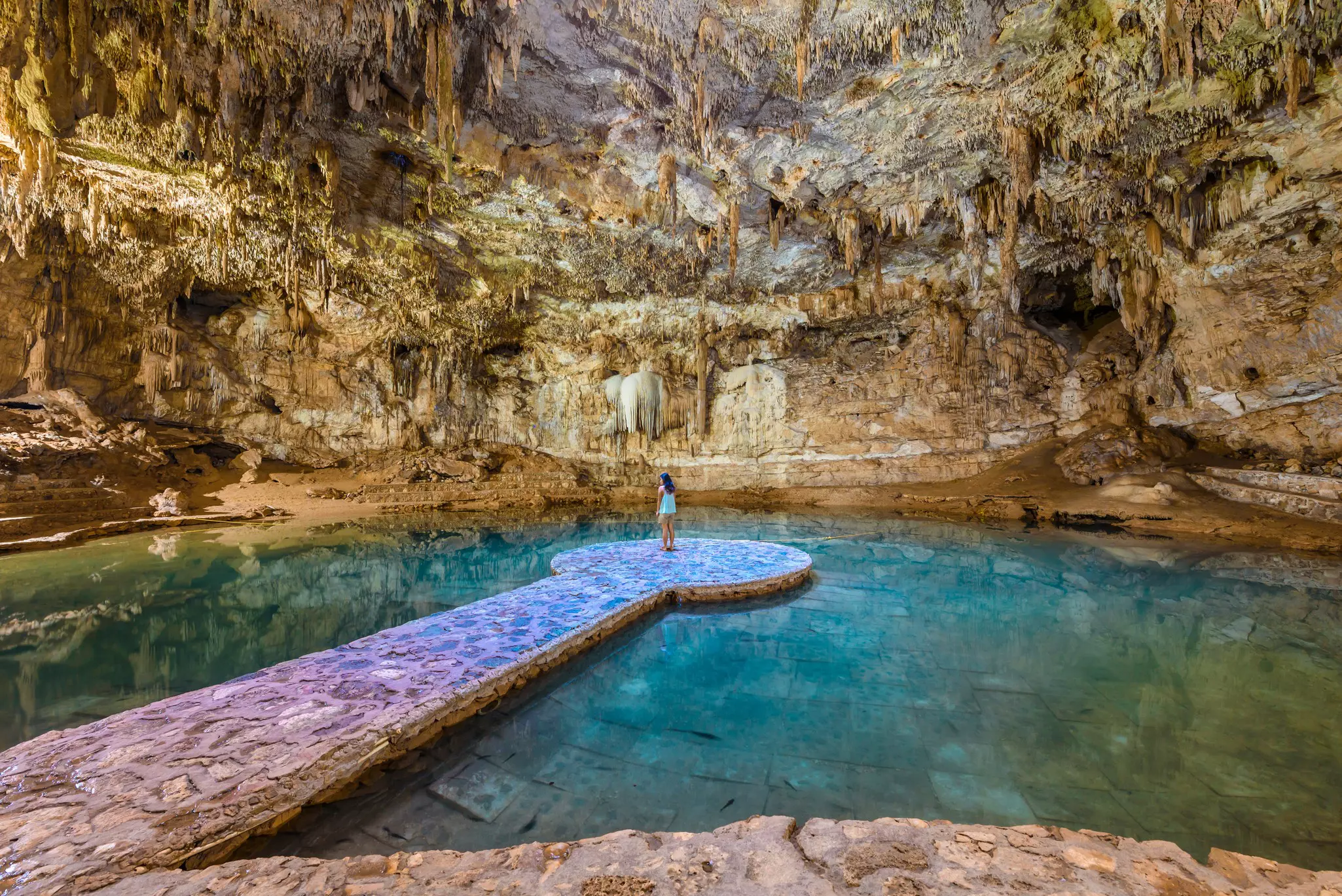 A woman stands on a platform in an underground pool in a cavern. Stalactites dangle from the high roof of the cavern.