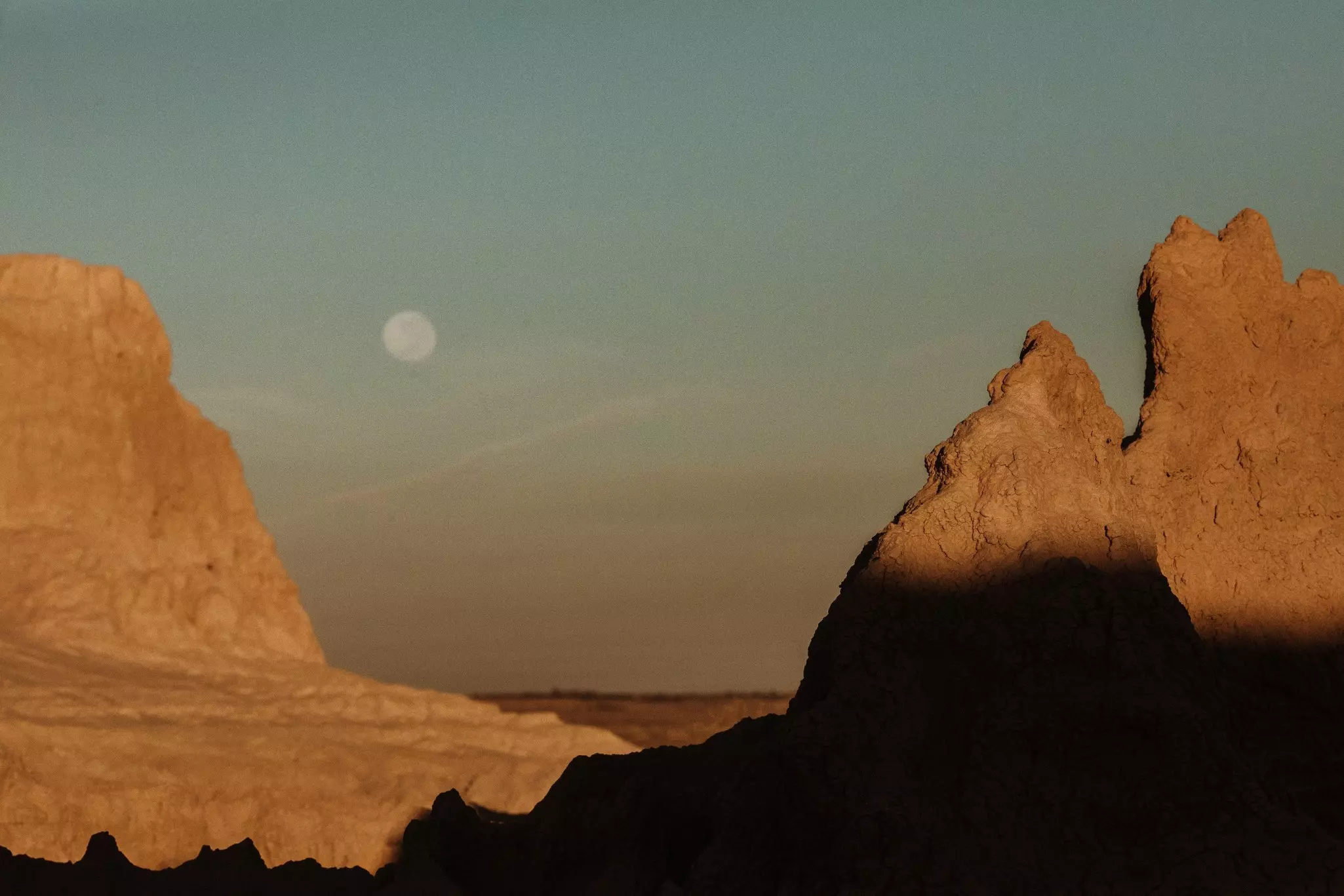 The moon in a pale blue sky overlooking two large rock mesas