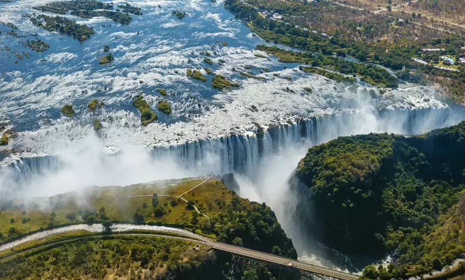 Aerial view of Victoria Falls plunging into a gorge on the Zambezi River, with mist, forest and the Victoria Falls Bridge