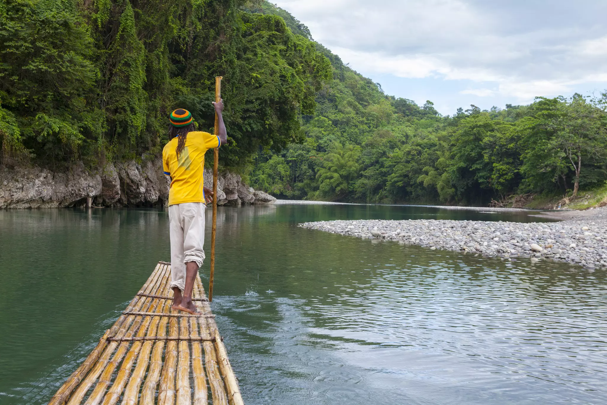 A man in a yellow shirt and striped hat stands at the front of a bamboo raft, pushing it forward through water with a long pole in the water; the waterway is lined by lush green vegetation, and a rocky shore is to the right.