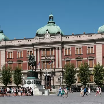 Red-brick and cream facade under three green domes, a bronze horseman on a plinth and people crossing the square