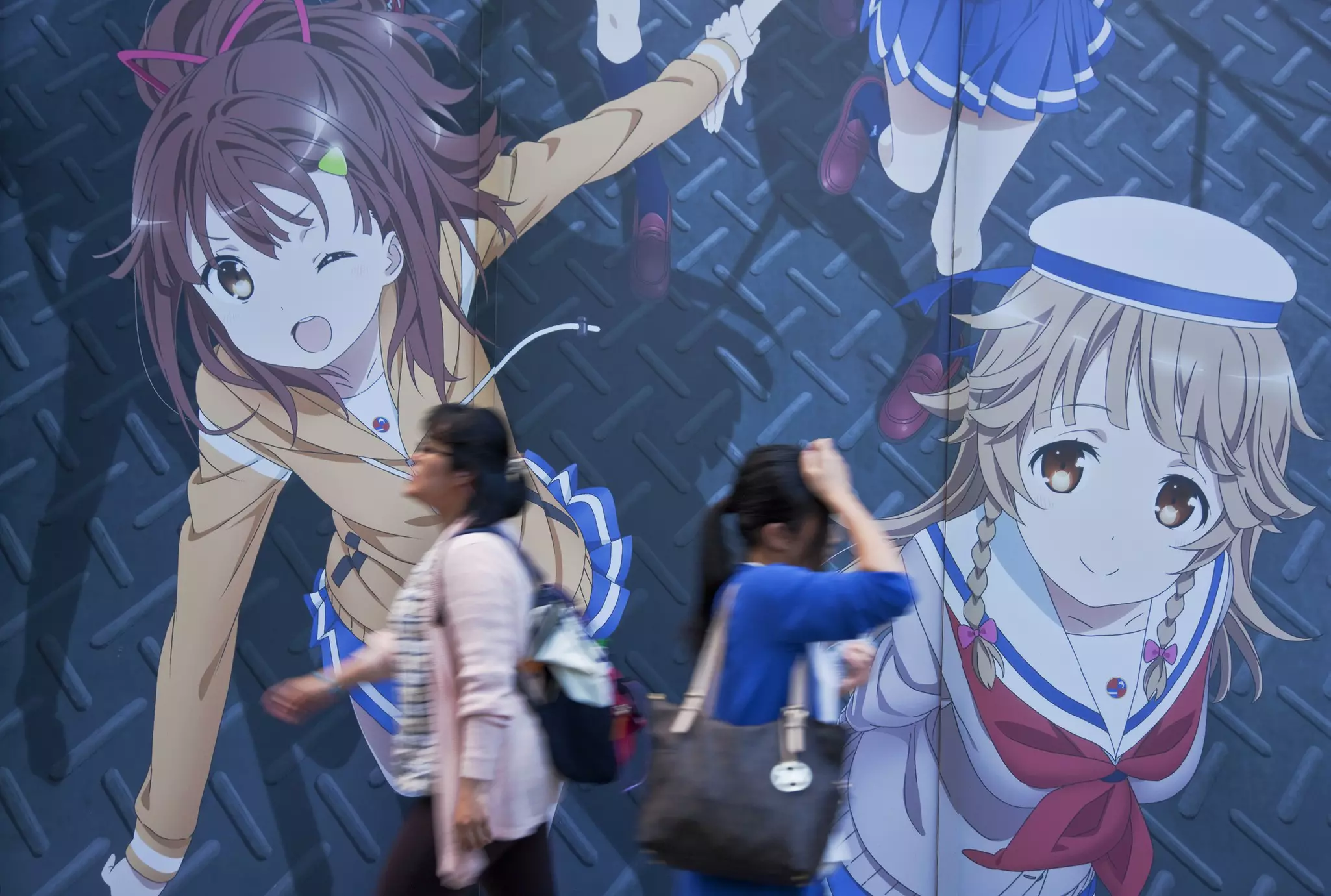 Two women walking in opposite directions pass a wall-sized anime mural