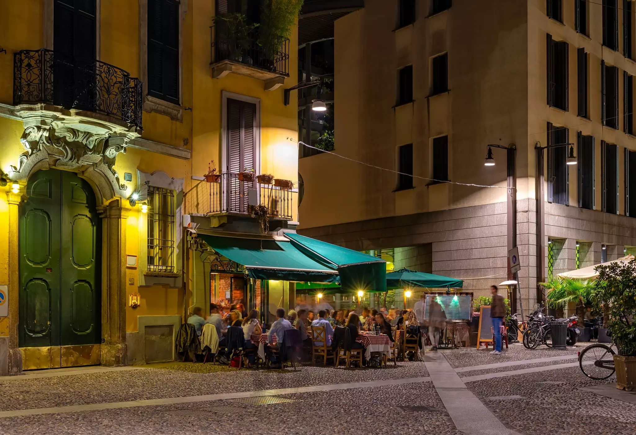 A view of a city street at night, with people enjoying a meal at tables on set up on the sidewalk.