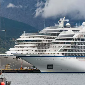 Large white cruise ships, including the Viking Jupiter, docked in a harbor beneath cloud-shrouded green mountains, a sailboat mast in front.