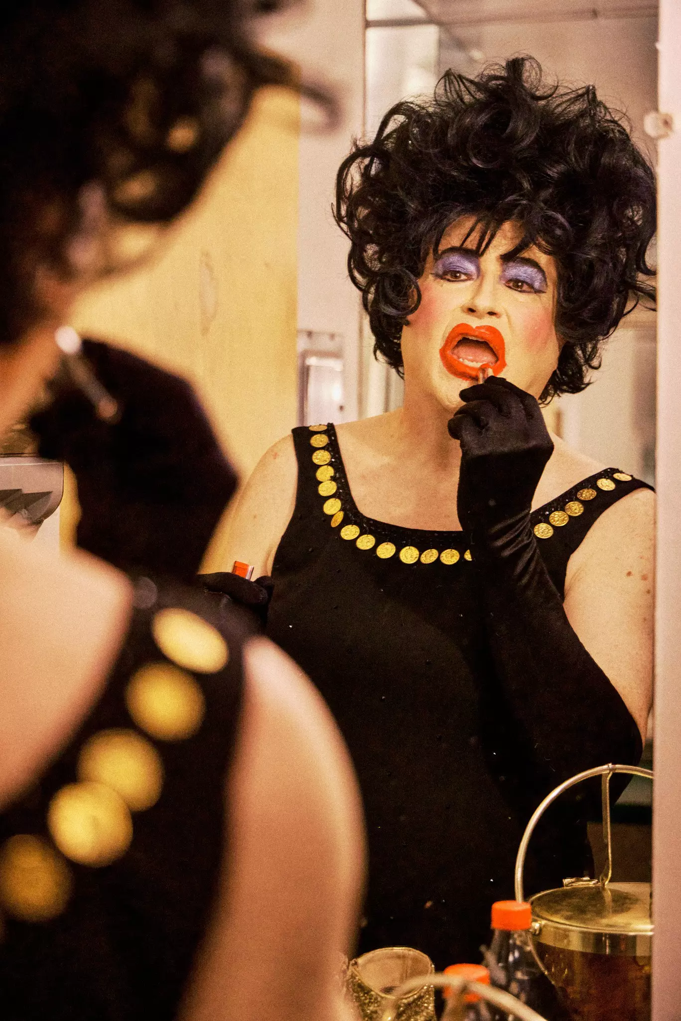 A drag queen in a black dress and a wig applies make up in a mirror reflection