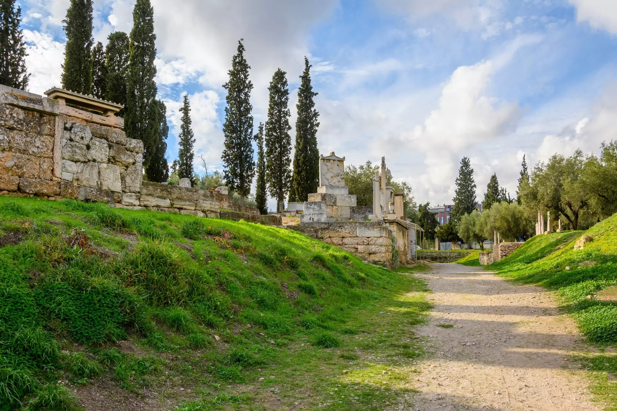 A pathway through a graveyard, with the ruins of ancient tombs rising on the hillside beside the path: Kerameikos, Athens