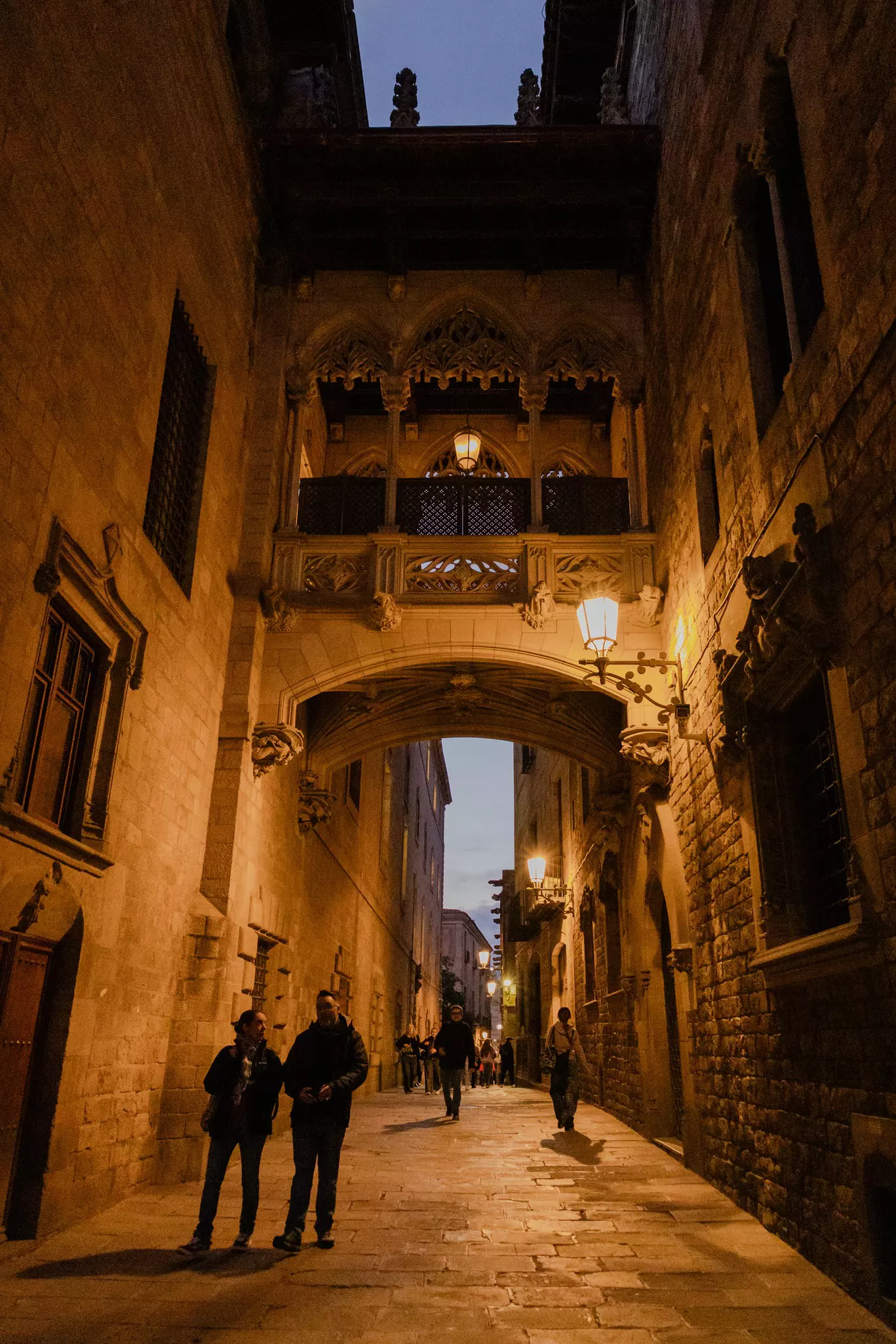 A Gothic bridge connects two buildings above an alleyway at dusk.