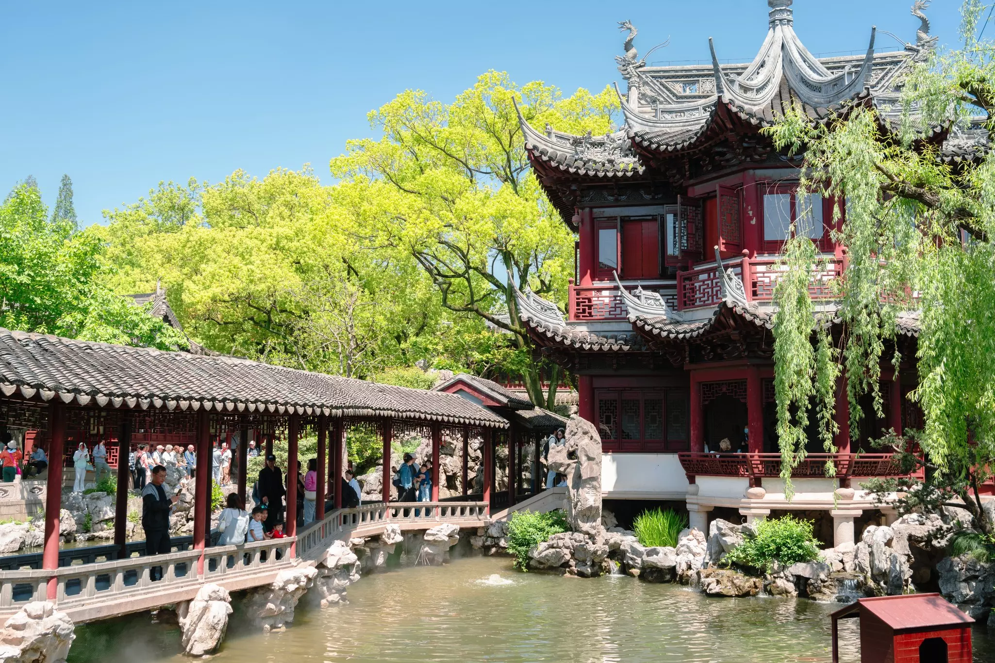 A garden with an ornate bridge over a pond leading towards a Chinese-style pagoda near a weeping willow tree.