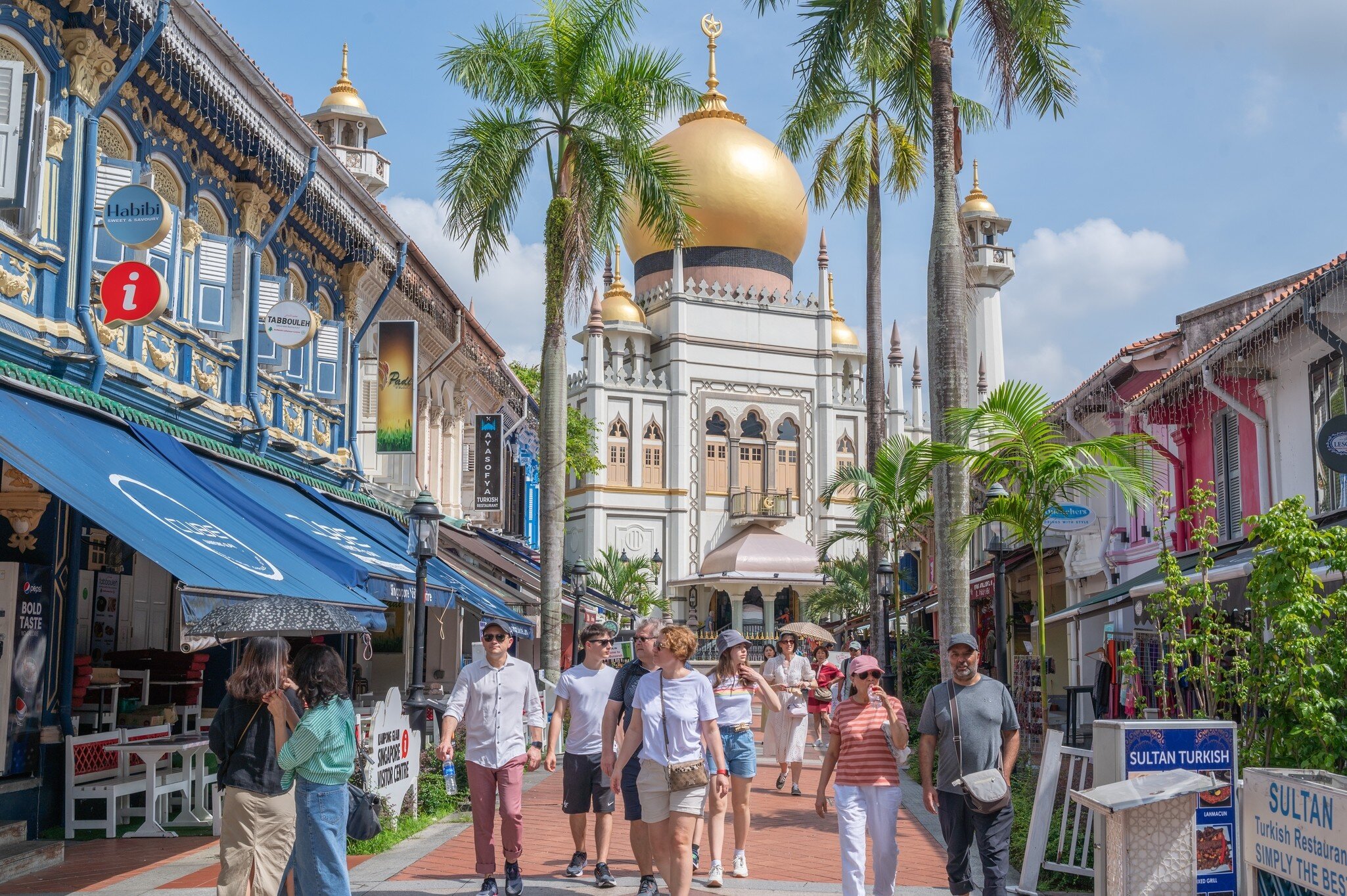 Visitors stroll a palm-lined street of colourful shophouses toward a white mosque topped by a large gold onion dome