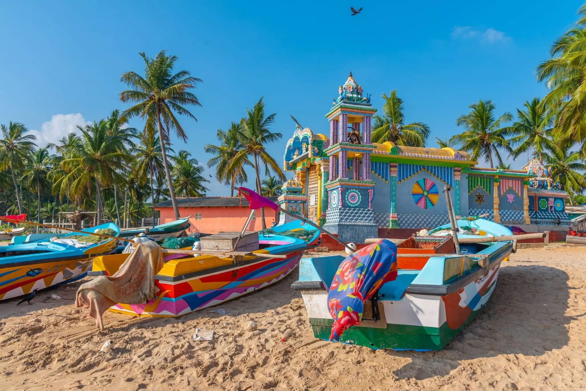 A row of brightly colored wooden boats stand on a sandy beach in front of a colorful temple 