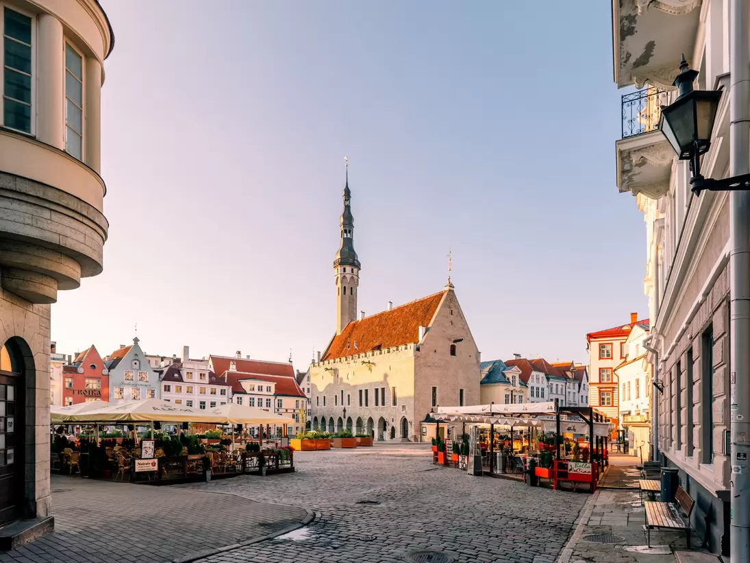 Cobbled square in soft morning light with a stone hall and slender spire, pastel gabled houses and cafés