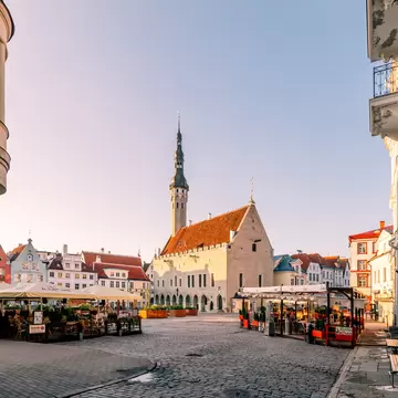 Cobbled square in soft morning light with a stone hall and slender spire, pastel gabled houses and cafés