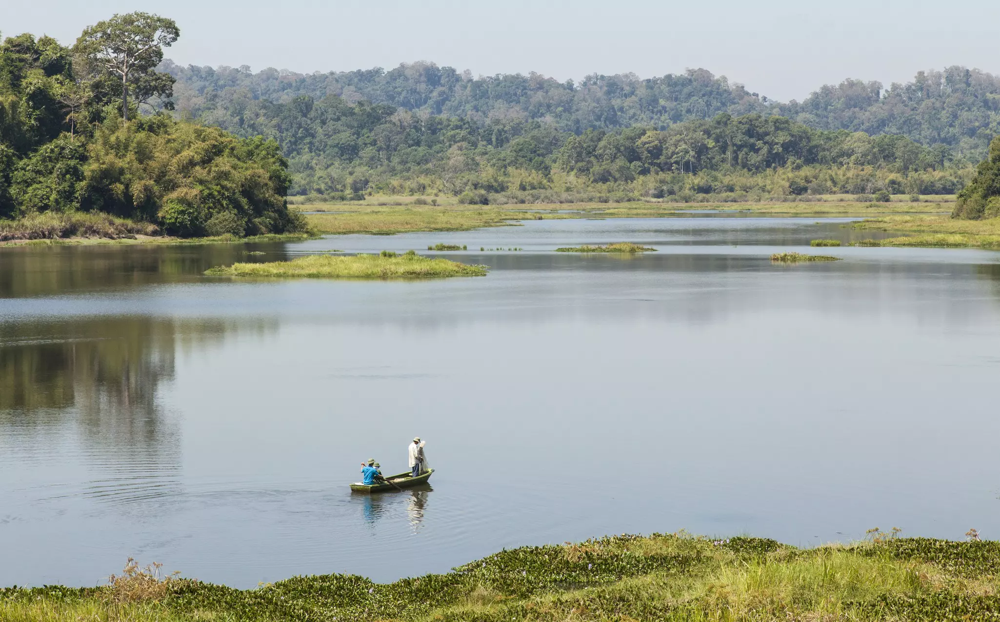 A small wooden boat containing two fishermen floats on a vast lake surrounded by green mountains in Cat Tien National Park, Vietnam.