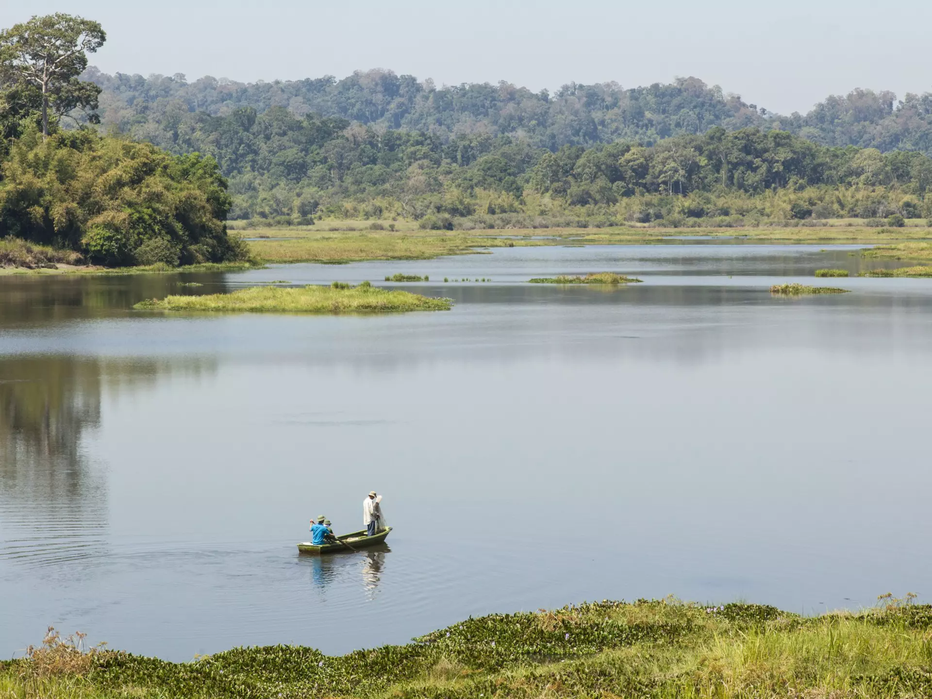 A small wooden boat containing two fishermen floats on a vast lake surrounded by green mountains in Cat Tien National Park, Vietnam.