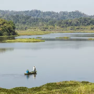 A small wooden boat containing two fishermen floats on a vast lake surrounded by green mountains in Cat Tien National Park, Vietnam.