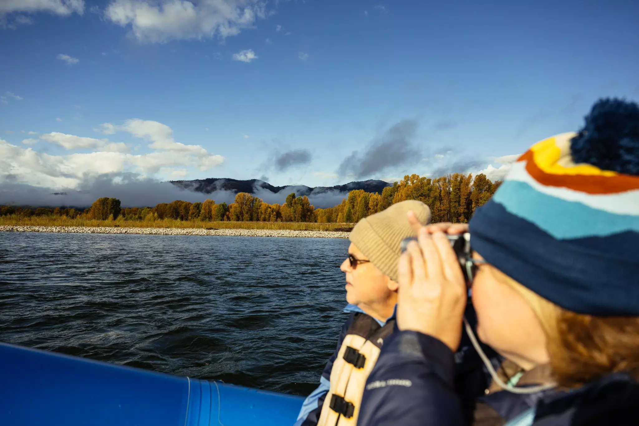 Two people in winter hats, one with binoculars, look out into river water under a nearly clear blue sky. 