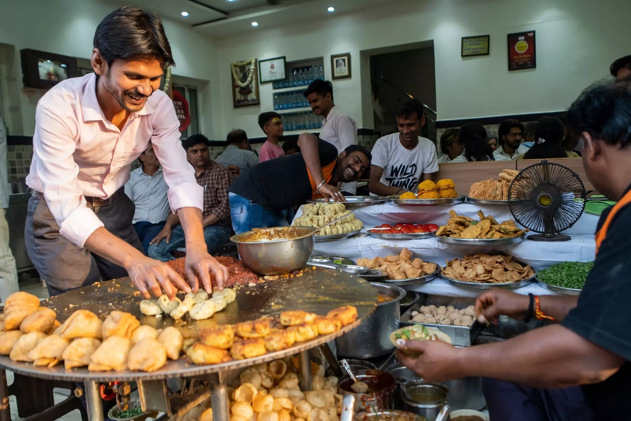 A man leans over a platter piled with fried snacks, as other vendors and diners look on in a dining room.