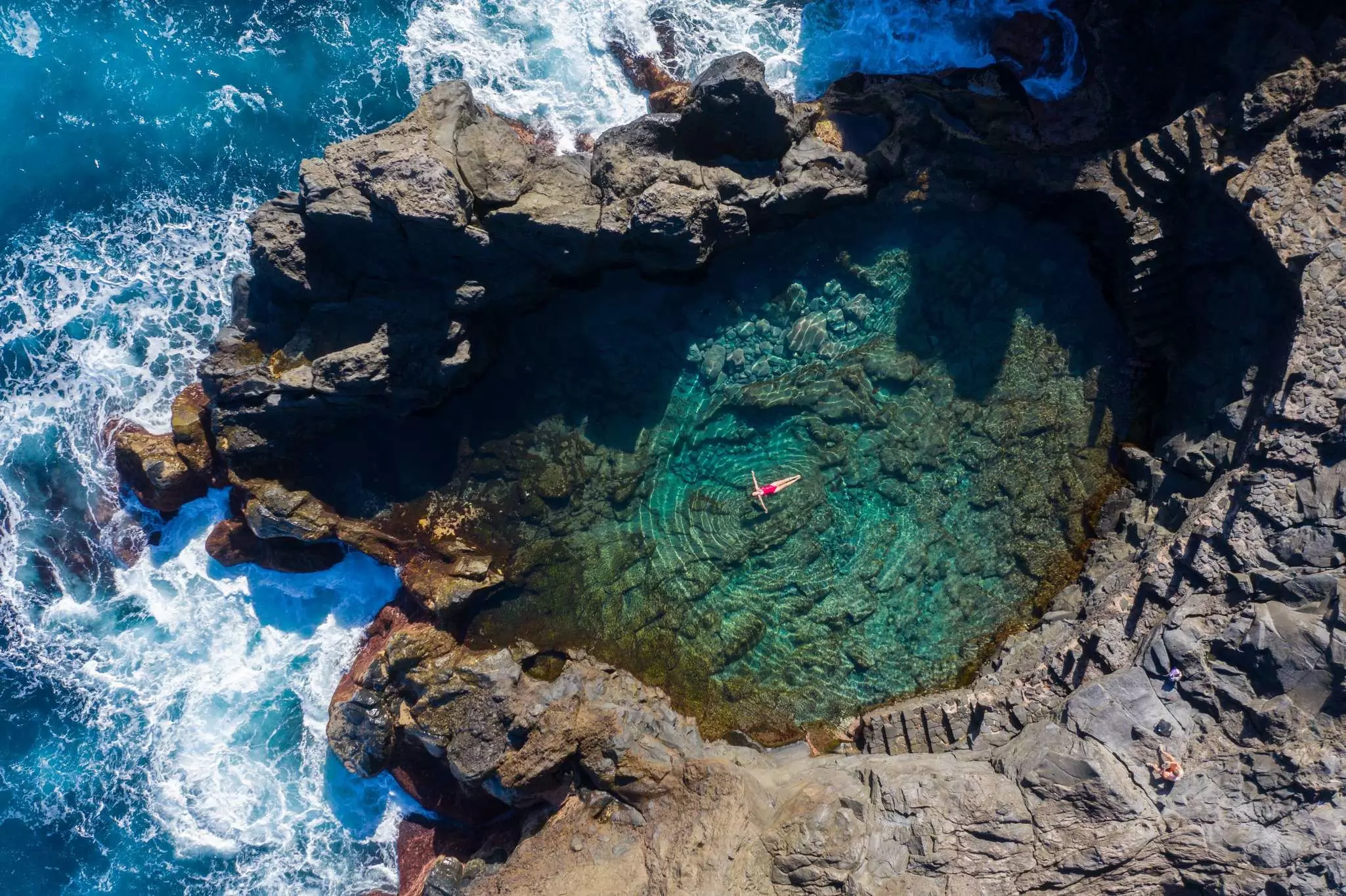 A woman in a red bathing suit floats in a large rock pool next to the crashing surf on a sunny day.