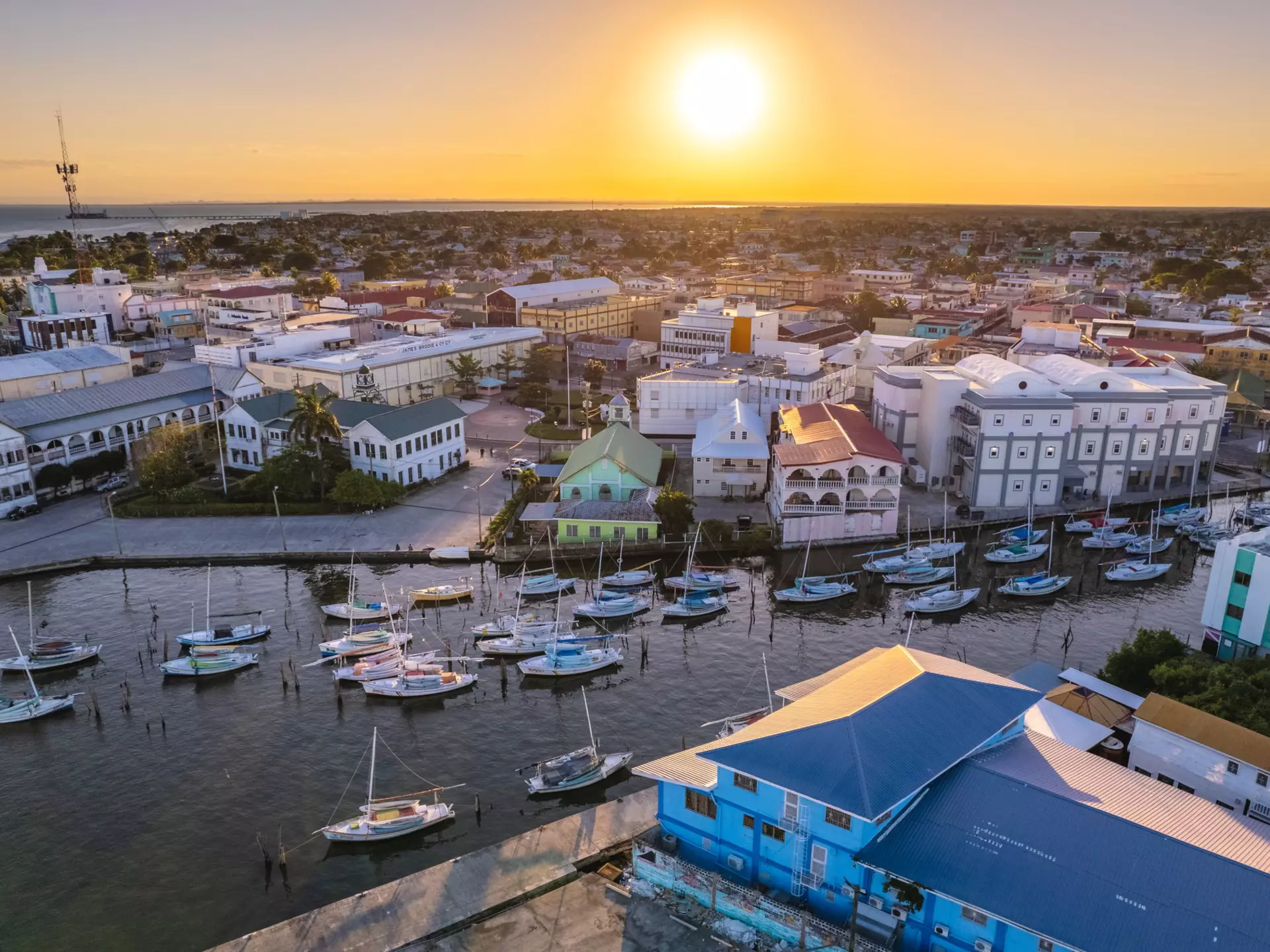 An aerial view of a waterfront in a city at sunset. Buildings with porches line the waterfront, while sailboats are moored in the water.