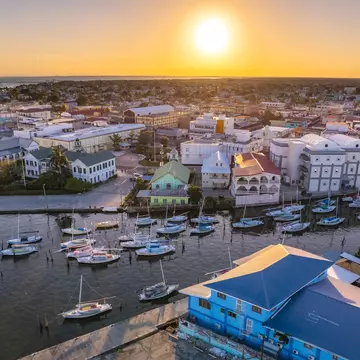 An aerial view of a waterfront in a city at sunset. Buildings with porches line the waterfront, while sailboats are moored in the water.