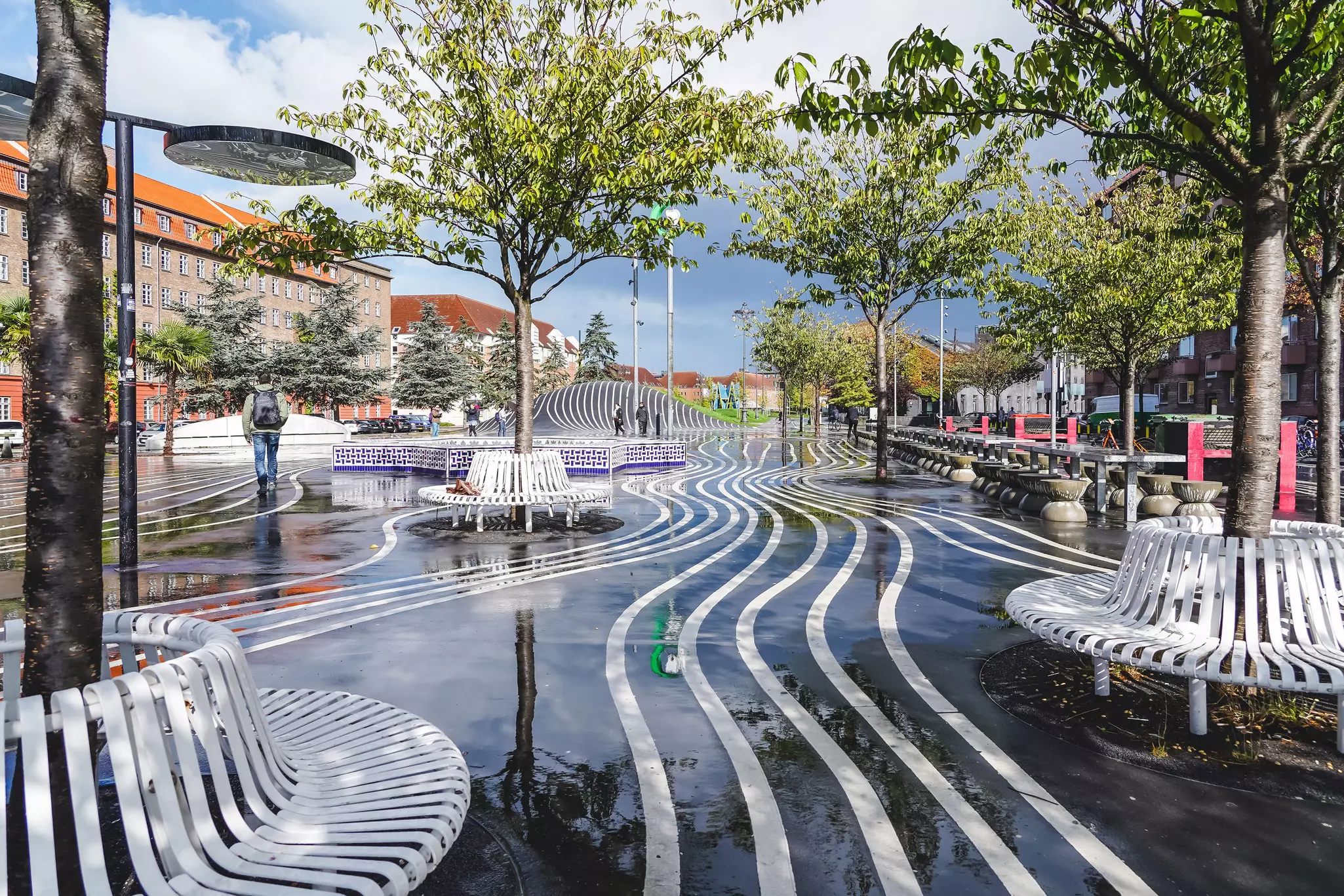 White stripes on dark pavement run in curving lines through an urban park; there are white circular benches surrounding trees, and the ground is wet, reflecting the trees and sky.