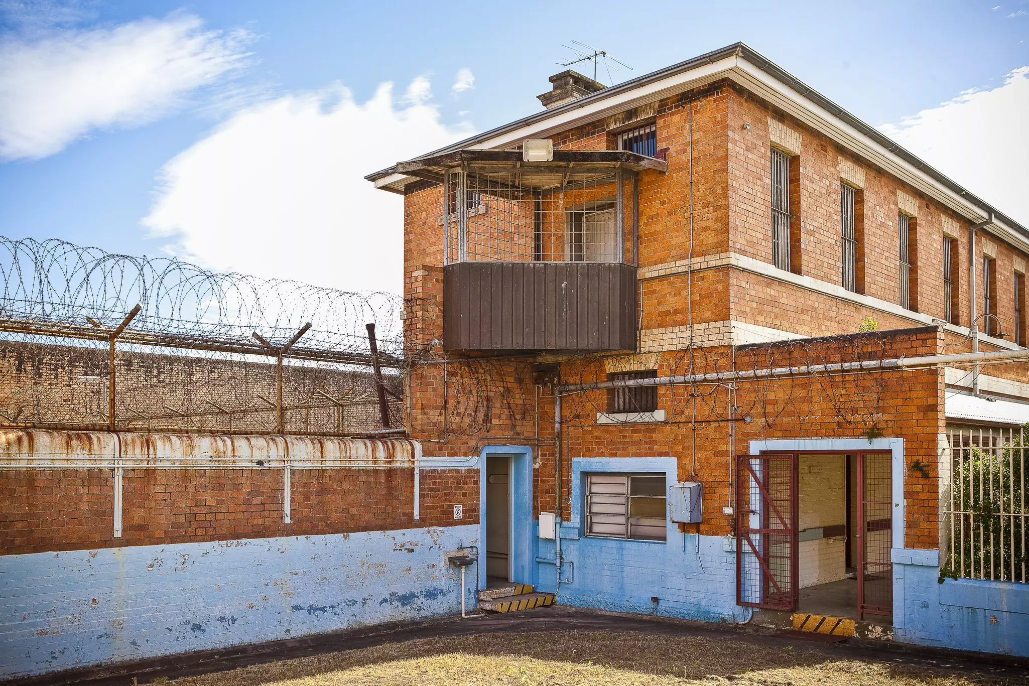 Outside of an old prison. The tall walls are fitted with barbed wire at the top; there is a door to the left and a fenced door to the right; haunted places world