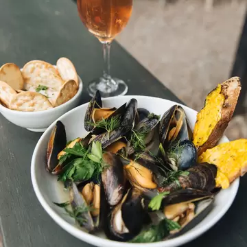 A white bowl of fresh mussels topped with green herbs and bread on the side. A glass of rosé and a small bowl of crackers sit behind 