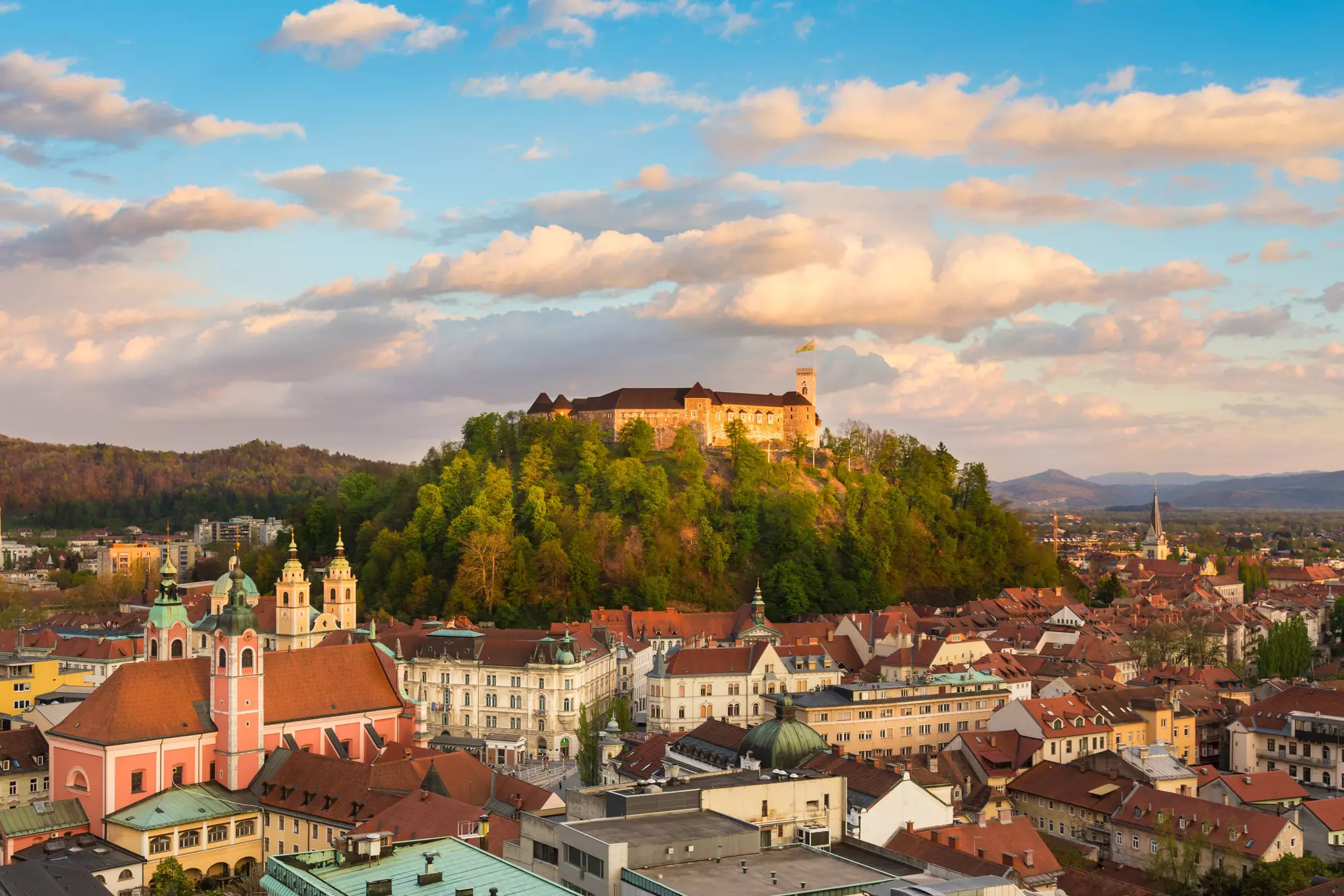 A large medieval castle sits on a hilltop surrounded by trees above a city.