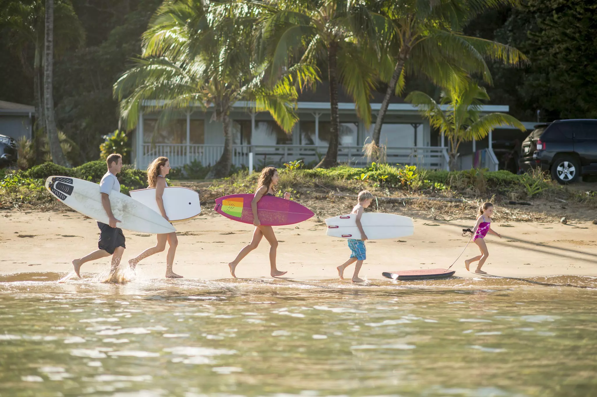 Adults and children carrying surfboards and boogie boards while walking down a beach in Hawaii