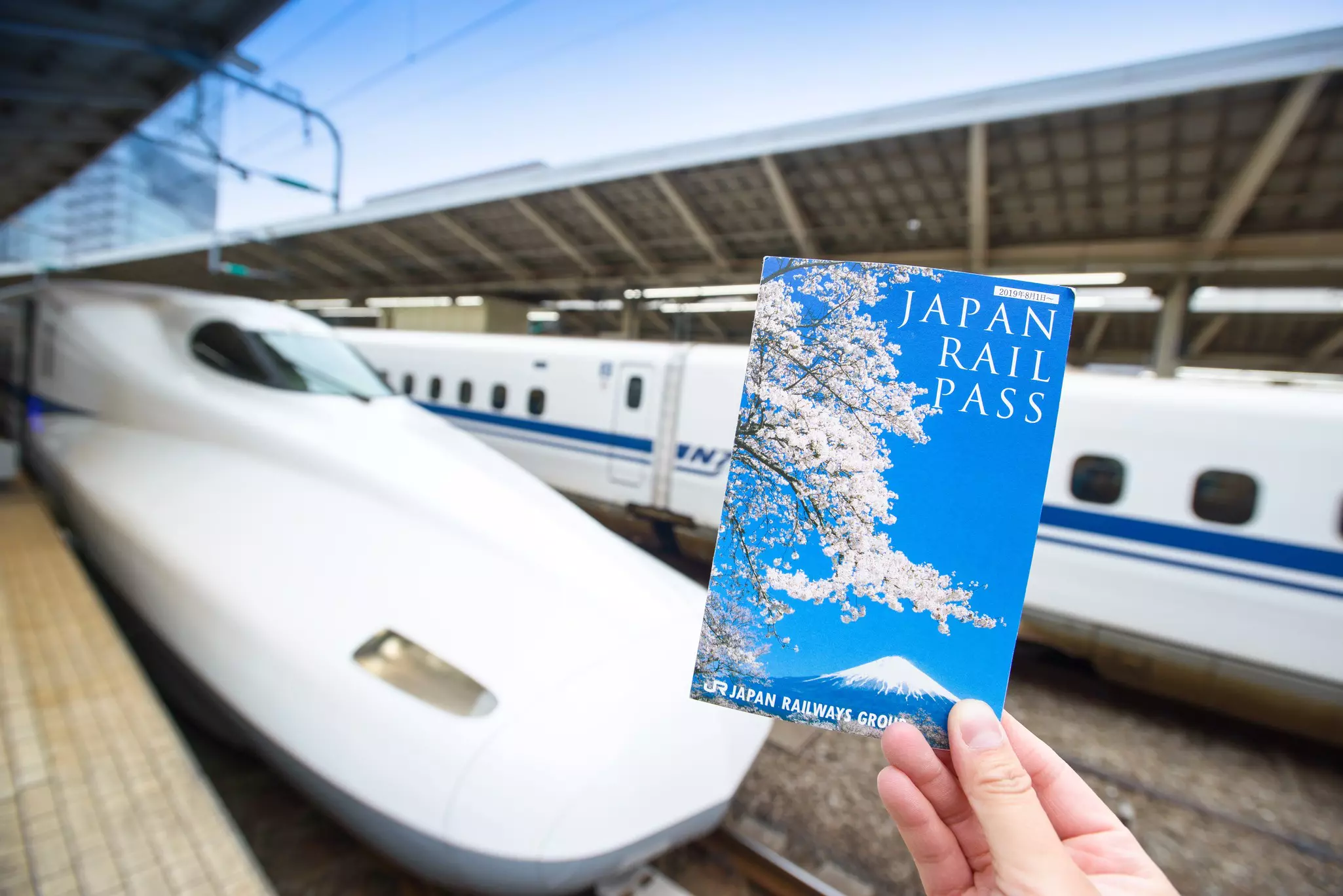 A hand holds up a Japan Rail Pass in front of the rounded nose of a bullet train at a station.