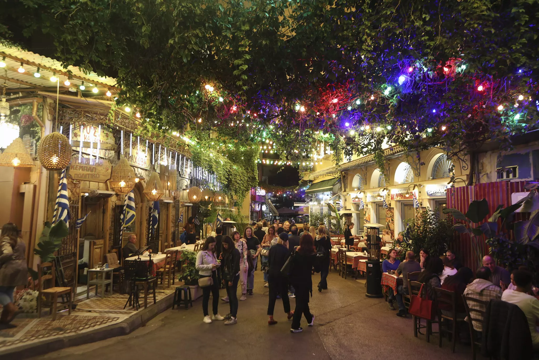 A group of people in a closed street with others sitting at outdoor café tables at nighttime with lights strung above.