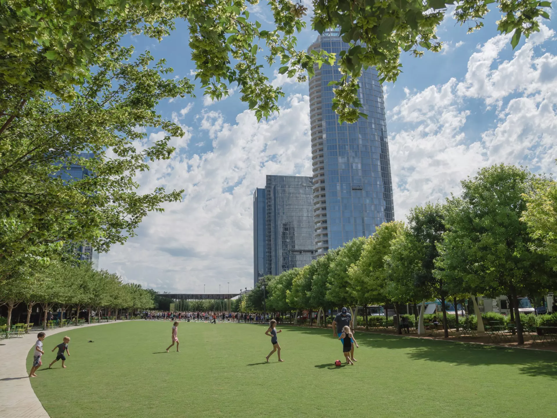 Children play soccer on a grass field in the shadow of tall buildings. 