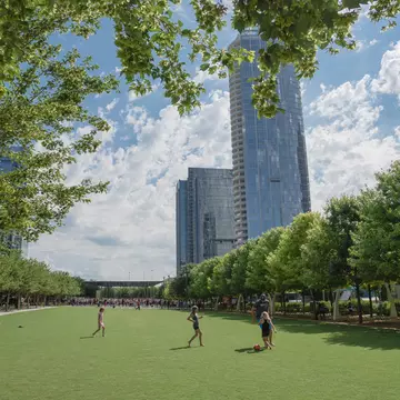 Children play soccer on a grass field in the shadow of tall buildings. 