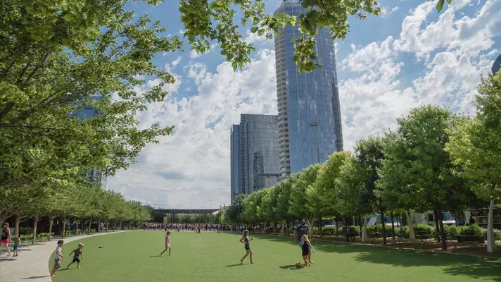 Green grass field surrounded by trees and park benches with children playing soccer 