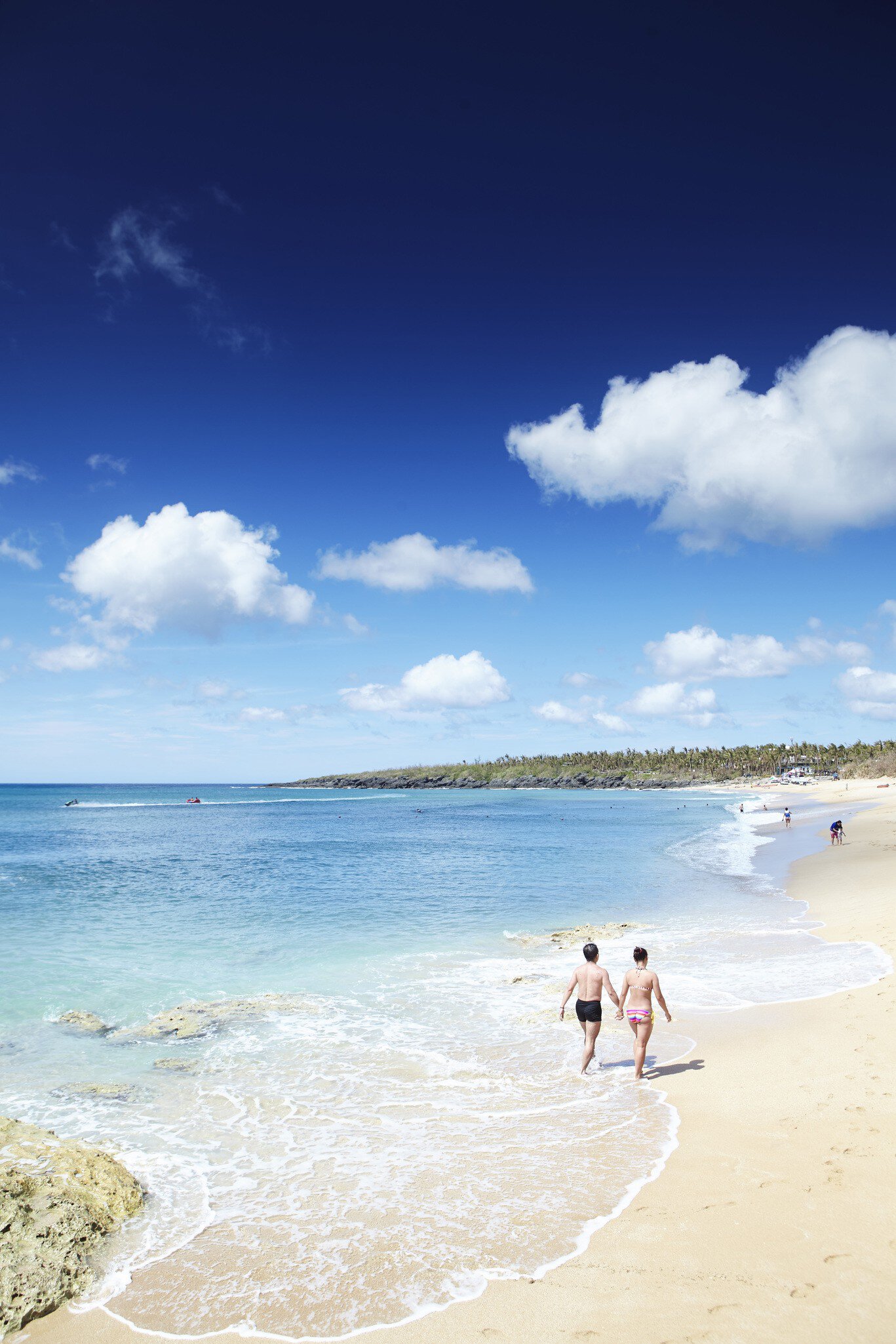 Two people in swimwear walking along the waterline of a pale sand beach, with turquoise sea and white clouds overhead.
