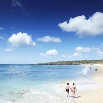 Two people in swimwear walking along the waterline of a pale sand beach, with turquoise sea and white clouds overhead.