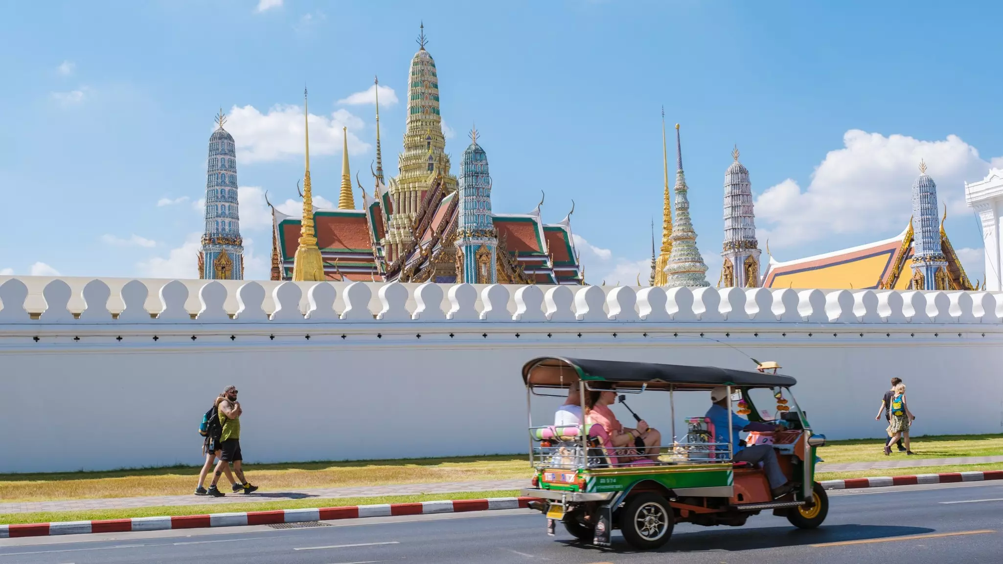 Passengers ride in a small motorized vehicle passing a grand palace with a decorative roof.