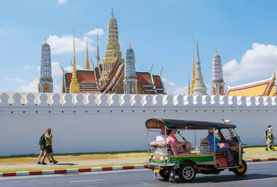 Bangkok Thailand 10 January 2025, colorful tuk-tuks glide past the stunning temple Wat Phra Kaew, east side of Grand Royal Palace under a bright blue sky. Tourists wander the vibrant streets