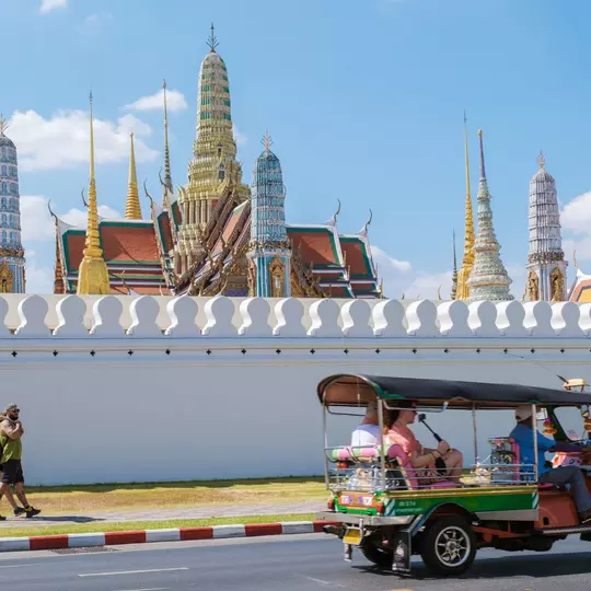 Bangkok Thailand 10 January 2025, colorful tuk-tuks glide past the stunning temple Wat Phra Kaew, east side of Grand Royal Palace under a bright blue sky. Tourists wander the vibrant streets