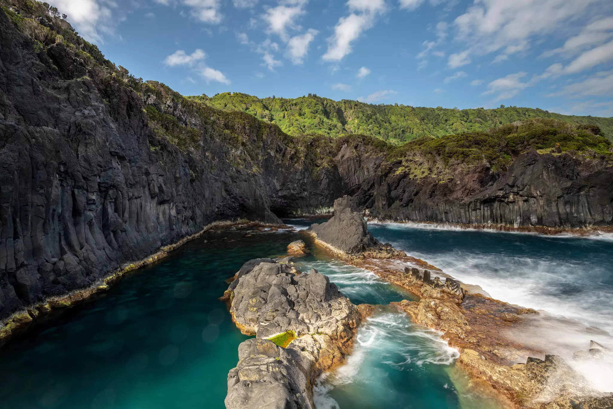 A pool formed by seawater having eroded volcanic cliffs. The contrast between the calm water of the pool and the white surf on the other side of the rocks is apparent.