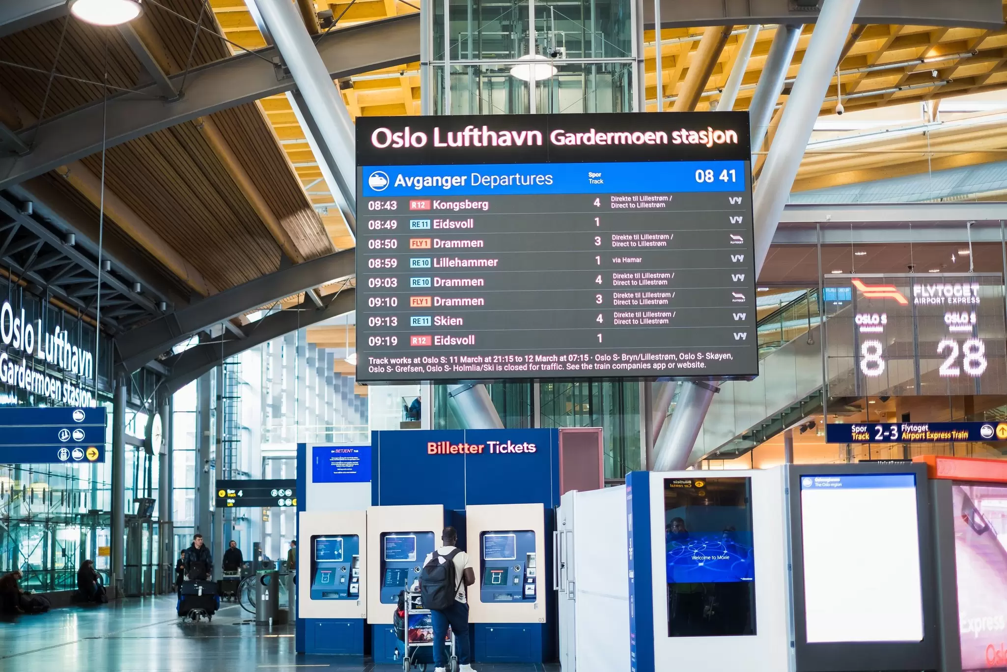 People with luggage in an airport where a large sign shows the train departures: Oslo, Norway