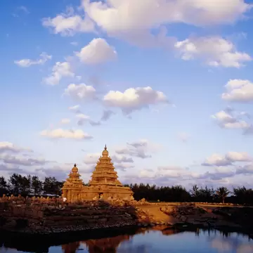 The stepped granite towers of Mamallapuram's Shore Temple in golden evening light, mirrored in dark water below a blue sky.