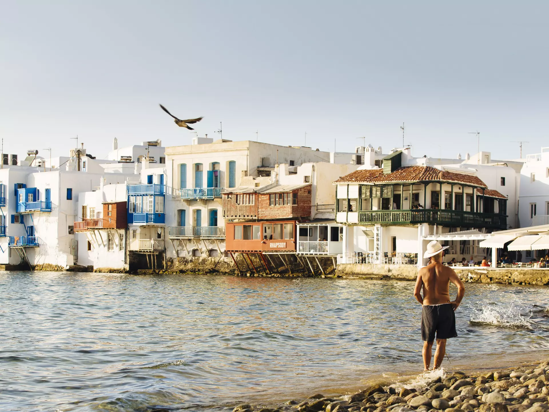 A man in shorts and a hat stands on the shore facing a row of beautiful buildings in Mykonos, Greece, as the evening light begins to fade. 